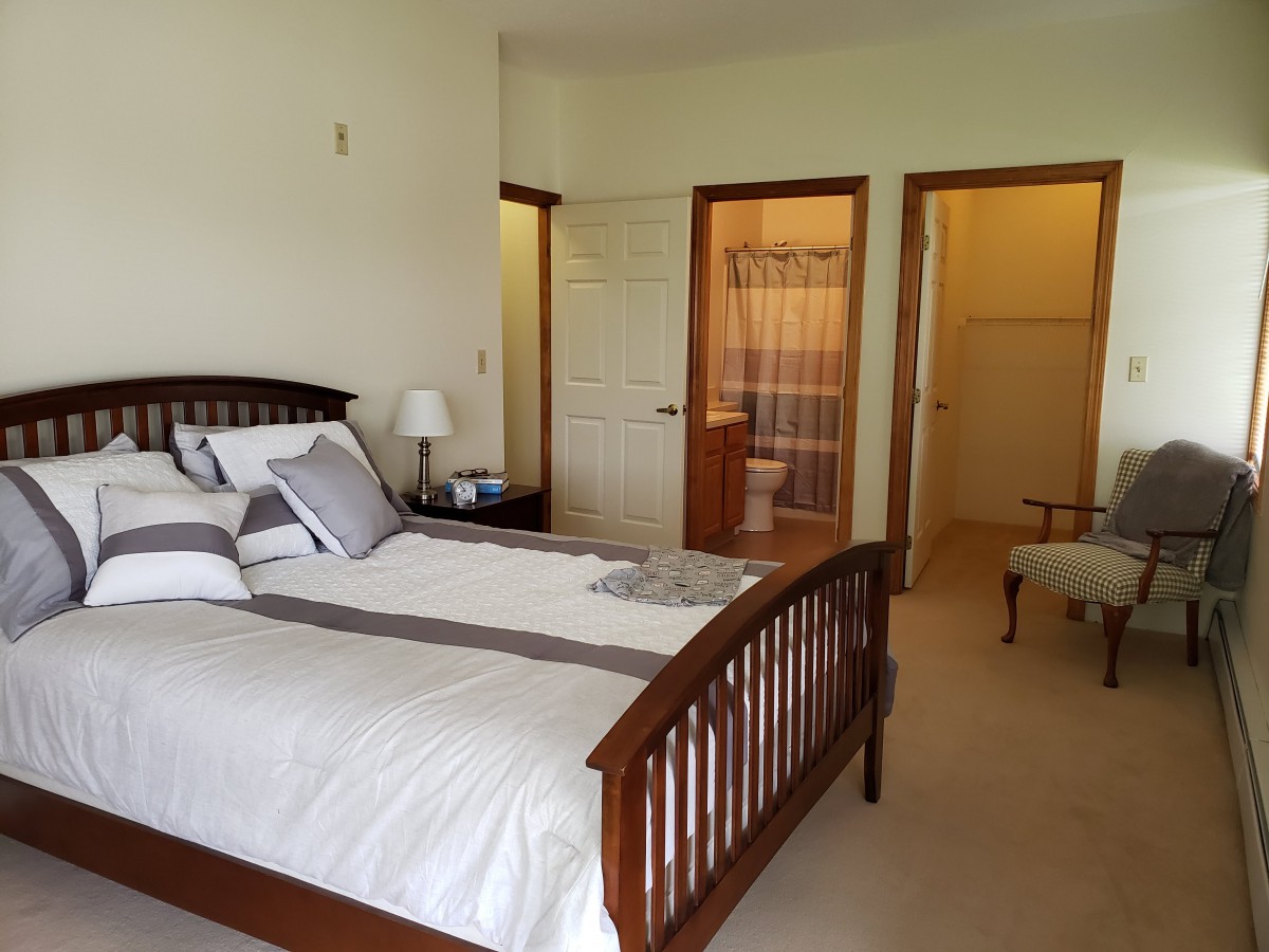 A cozy bedroom featuring a wooden bed frame, neatly arranged pillows and a view into an adjoining bathroom.