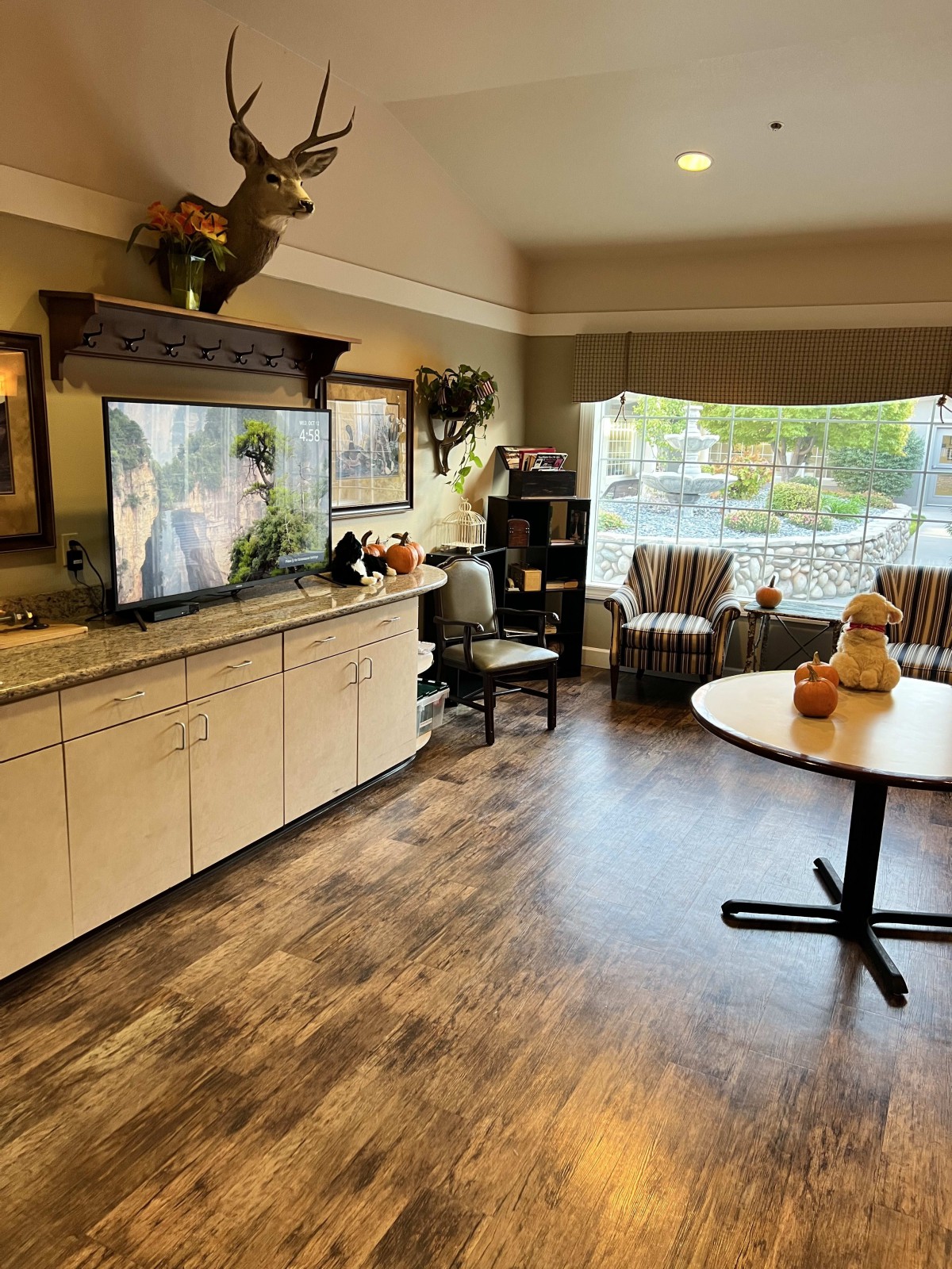 A cozy living room with a large television, seating area, and a table near a bright window.