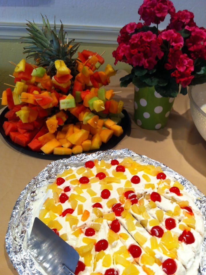 A dessert table with a fruit-topped cake and colorful fruit arrangements next to a vase of flowers.