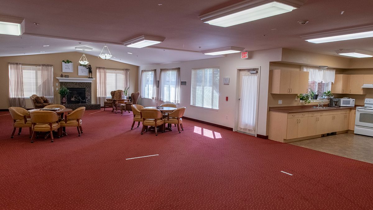 A spacious room with red carpet, featuring several tables and chairs, large windows, and overhead lighting.