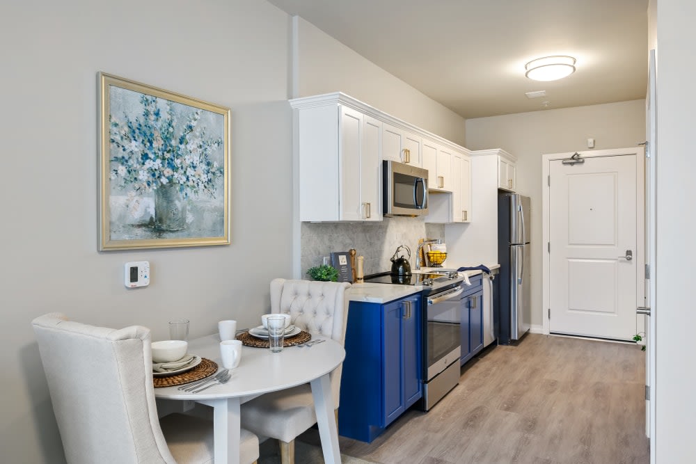 A modern kitchen with blue and white cabinets, a stainless steel stove and microwave, a small dining table set for two, and wood flooring.