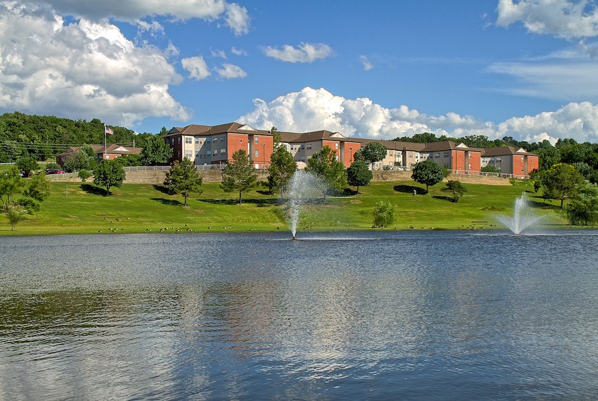 A serene view of a lake with a fountain in front of residential buildings and lush greenery under a bright blue sky.