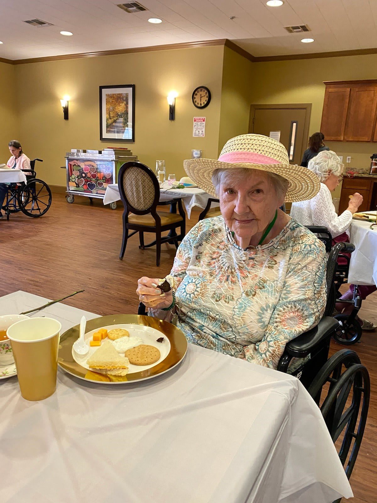 An elderly woman wearing a hat, sitting in a wheelchair and enjoying a meal in a dining area.