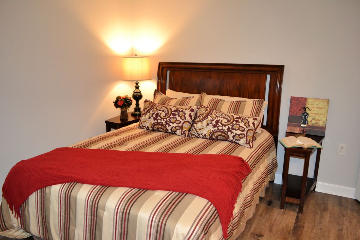 A cozy bedroom with a neatly made bed, featuring striped bedding and a bright red accent. A lamp on the nightstand provides warm lighting.