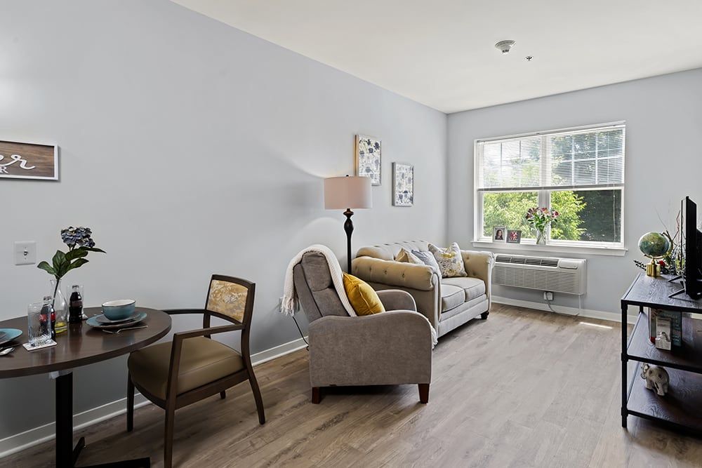 A cozy living room with a sofa and armchair, decorated in neutral tones and brightened by a window.
