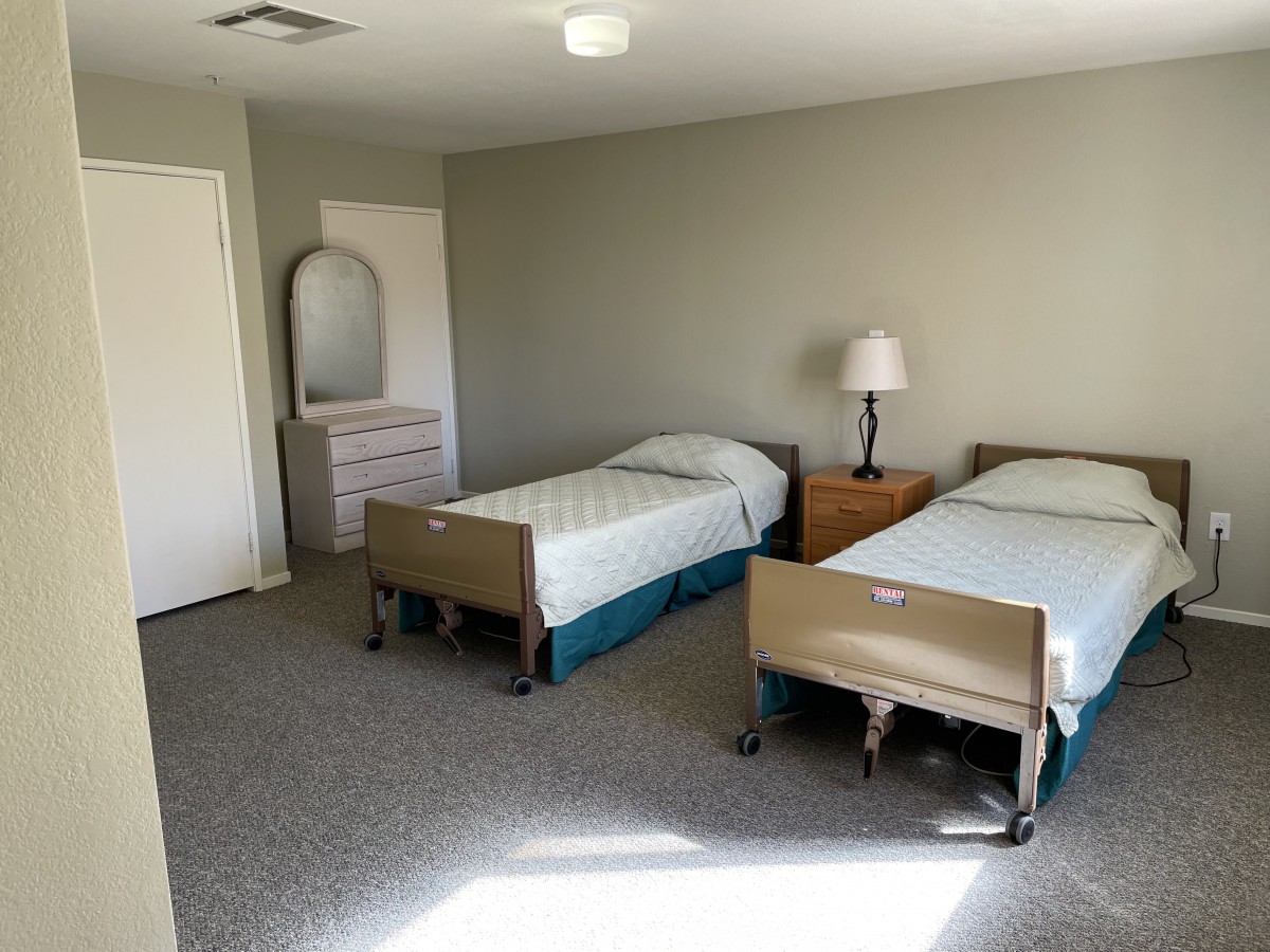 A minimalist bedroom with two hospital-style beds, a lamp, and a dresser against grey walls.