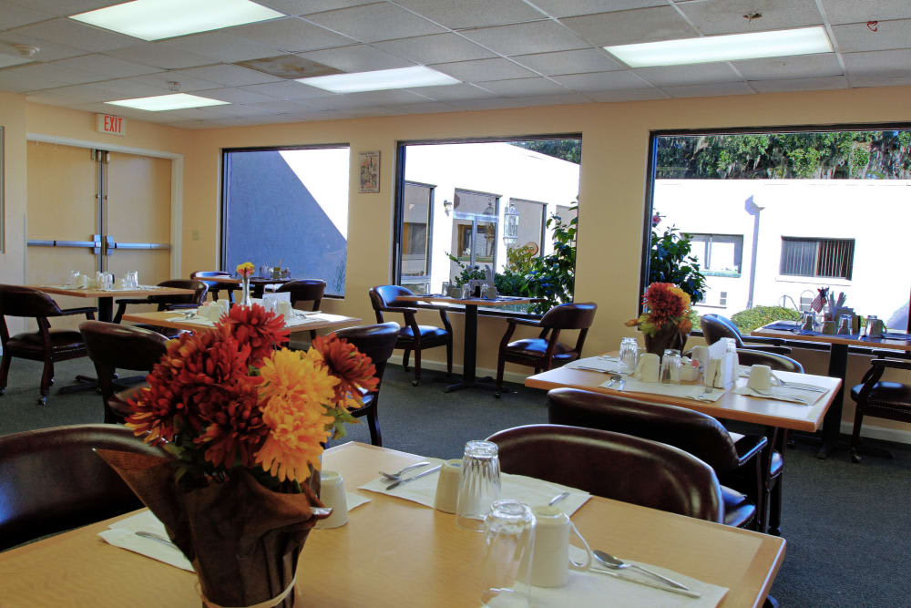 A cozy dining area with tables set for a meal, featuring floral centerpieces and ample natural light from large windows.