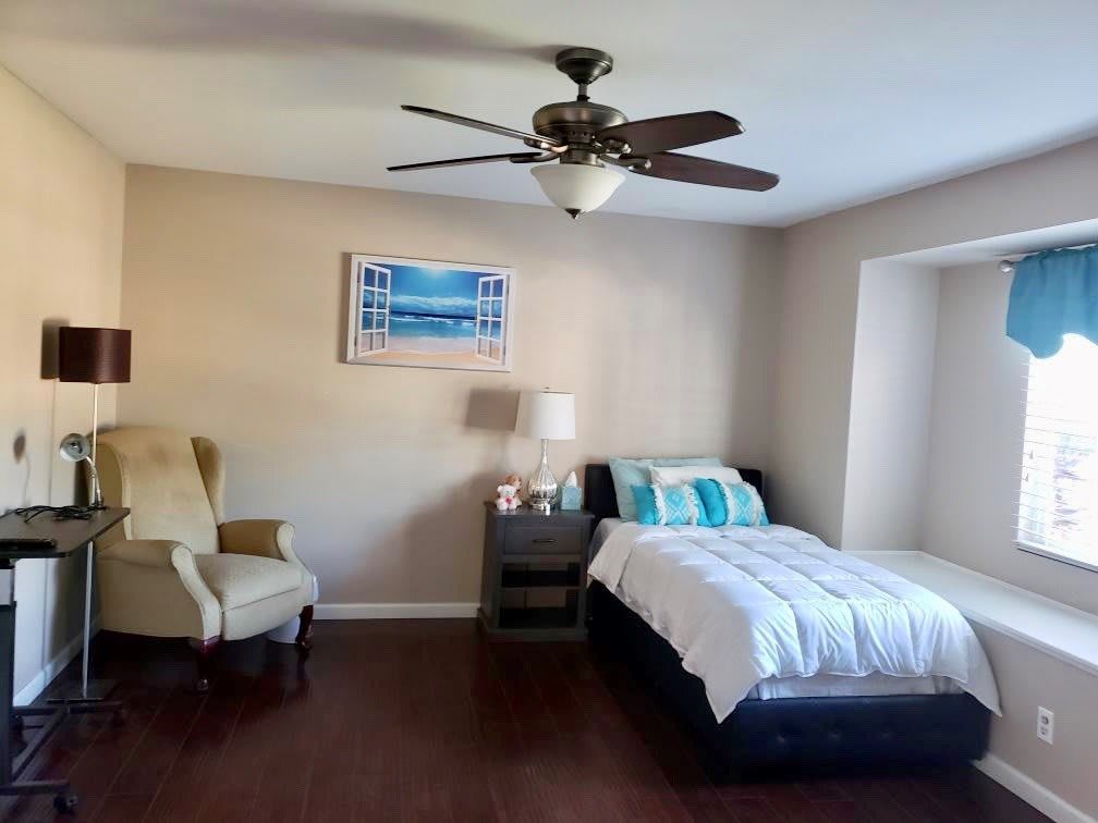 A modern bedroom features a bed, chair, nightstand with a lamp, and a ceiling fan.