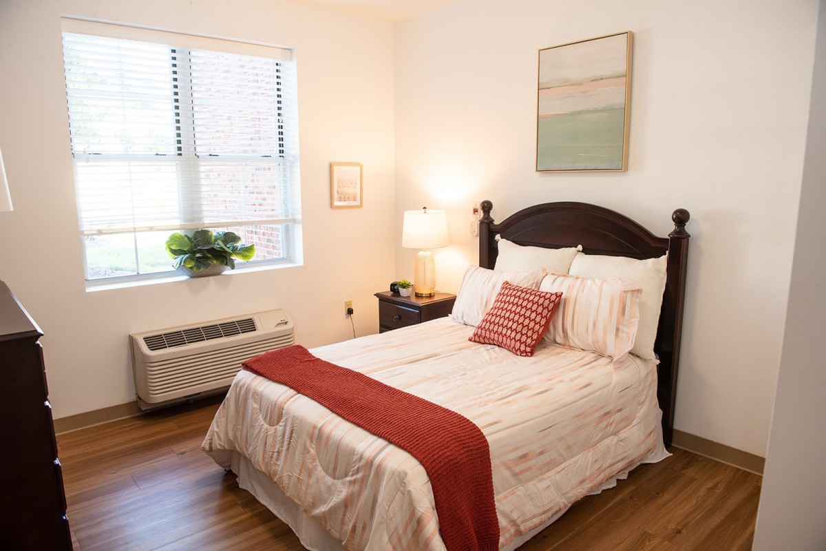 A cozy and well-lit bedroom featuring a neatly made bed with red accents, a bedside lamp, and artwork on the wall.