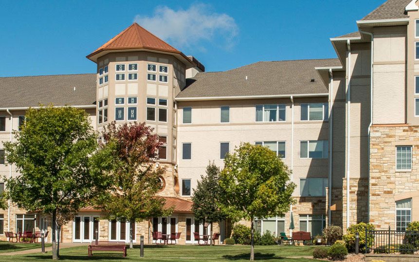 A multi-story residential building with a distinctive tower and surrounded by lush greenery and trees under a clear blue sky.