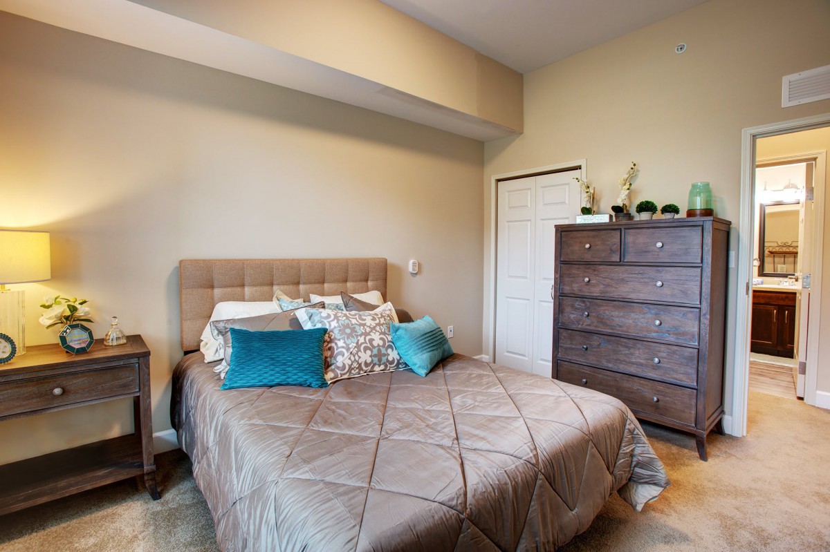 A cozy bedroom with a neatly made bed, colorful pillows, and a wooden dresser.
