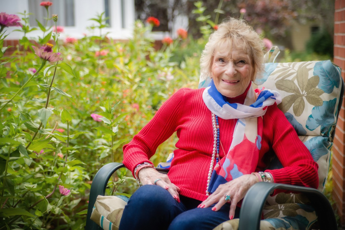 An elderly woman sitting comfortably in a garden, wearing a red sweater and colorful scarf.