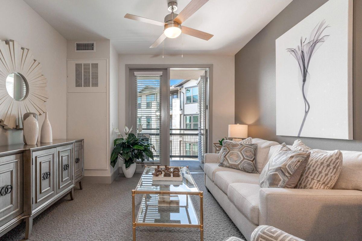 A cozy and well-decorated living room with a sofa, glass coffee table, and ceiling fan, featuring modern decor and ample natural light.