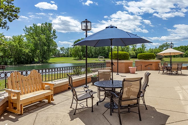 A scenic outdoor patio with a table, chairs, and umbrella overlooking a lake and trees.