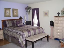 A cozy bedroom featuring a bed with purple accents, complemented by matching window curtains and decorative wall art.