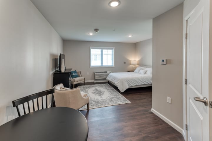 A modern bedroom with hardwood flooring, featuring a cozy bed, a lamp, and a window providing natural light.
