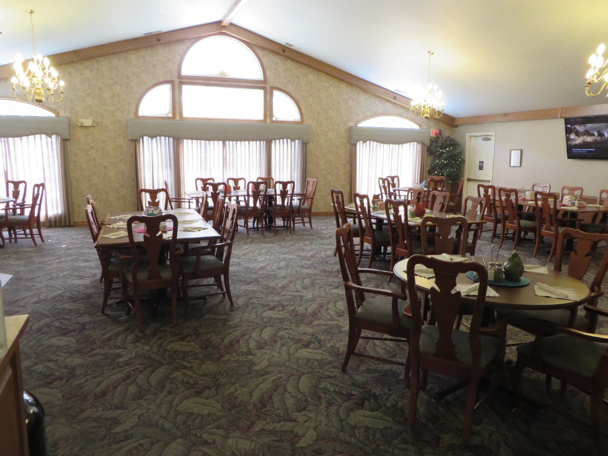 An empty dining room with multiple tables and chairs, featuring large windows and a carpeted floor.