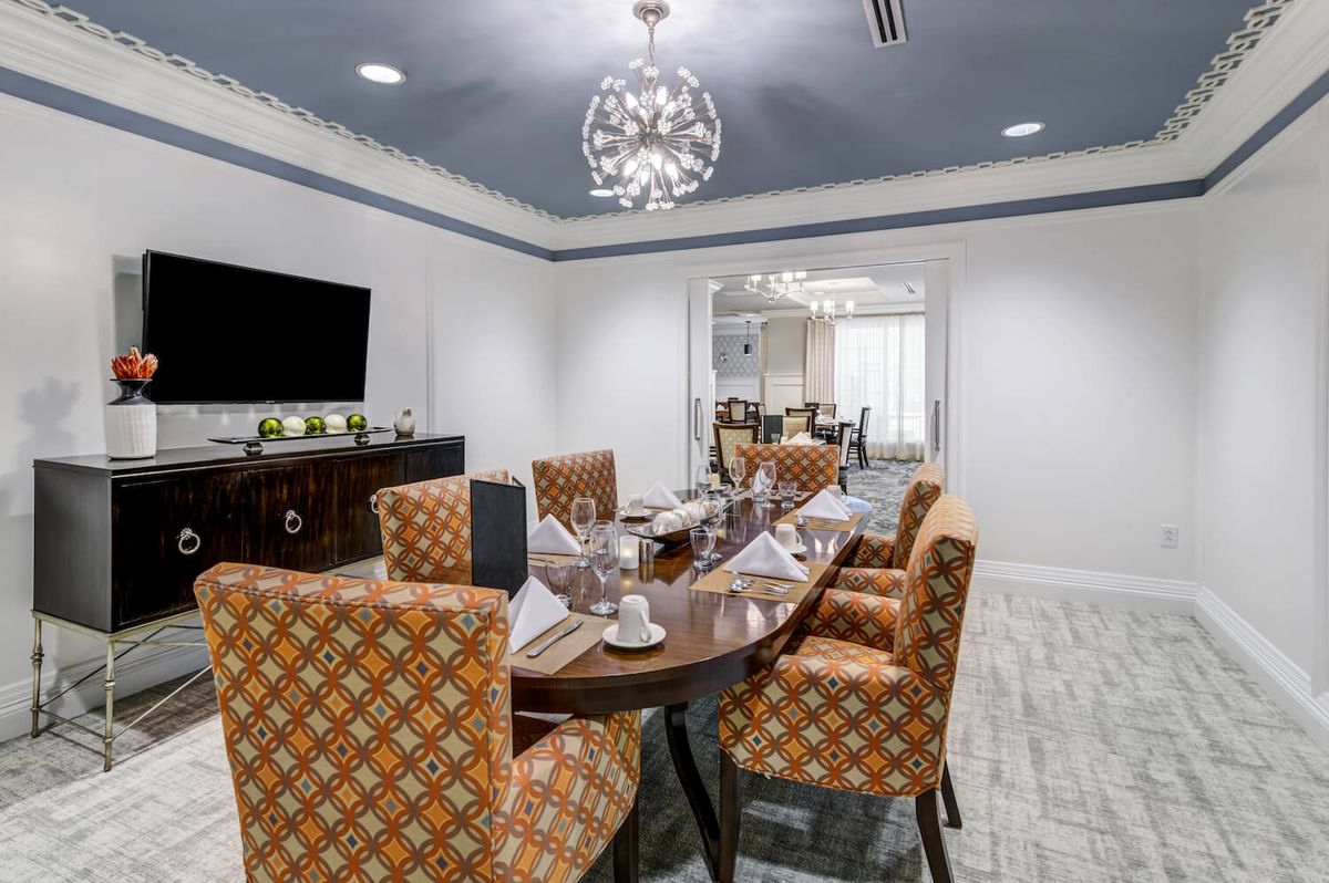 A modern dining or conference room featuring a large table with patterned chairs, a chandelier, and a flat-screen TV on the wall.