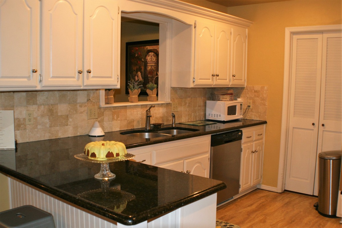A kitchen with white cabinets, a dark countertop, and various appliances, including a microwave and dishwasher. A cake is displayed on the counter.