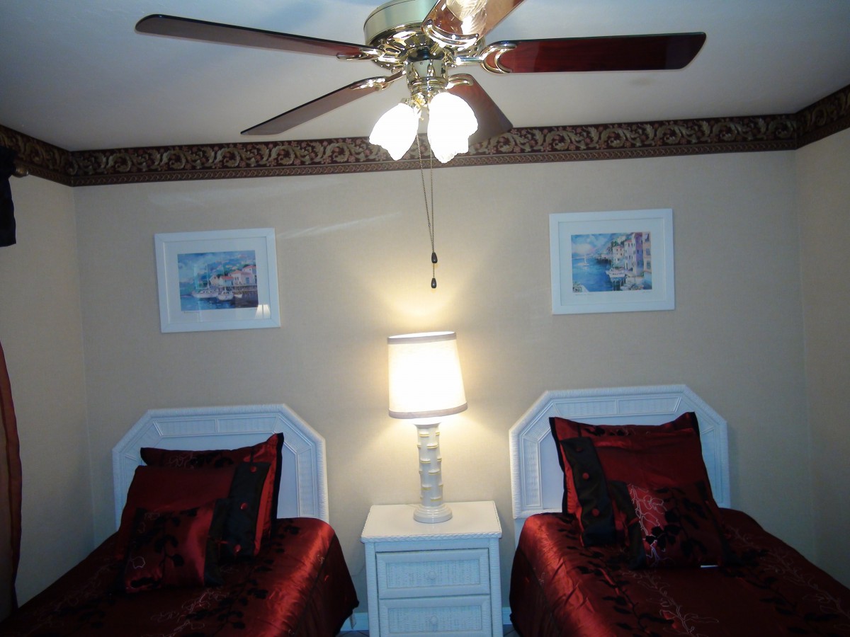 A cozy bedroom with twin beds featuring red bedding, a central nightstand with a lamp, and framed wall art beneath a ceiling fan.