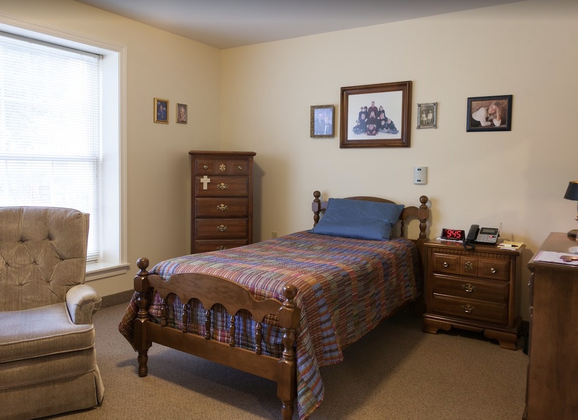 A cozy bedroom featuring a single wooden bed with a plaid comforter, accompanied by matching wooden furniture including a dresser and nightstand. The room is decorated with framed pictures on the walls and receives natural light from a large window.