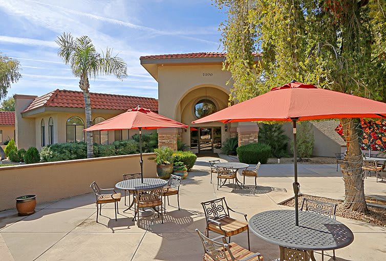 A sunny outdoor patio area with tables, chairs, and umbrellas near a building entrance surrounded by palm trees.