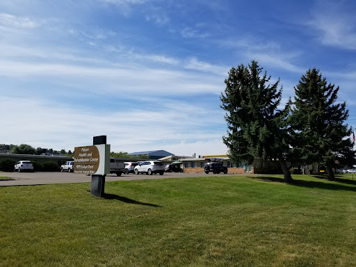 A sunny day view of a building with parked cars, surrounded by trees and open grass.