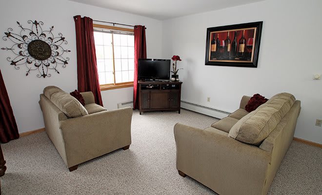 A cozy living room with beige sofas, a TV on a stand, and red curtains framing a large window.