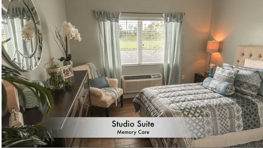 A cozy studio suite designed for memory care, featuring a comfortable bed, armchair, and well-arranged decor with a view of the outdoors.