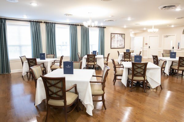 A neatly arranged dining room with tables set for guests, featuring elegant decor and large windows with blue curtains.