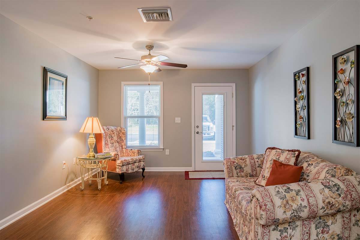 A cozy living room with wooden flooring, featuring a sofa, armchair, a lamp, and a ceiling fan.