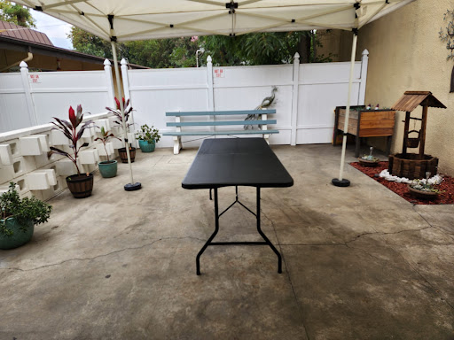A black folding table set up under a tent in an outdoor patio area, surrounded by potted plants and a white fence.