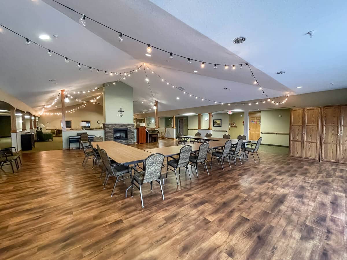 A spacious indoor meeting room with string lights, featuring rows of tables and chairs on a hardwood floor.
