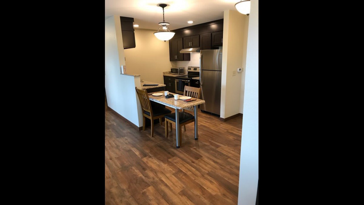 A modern kitchen with stainless steel appliances and a small dining area featuring wood flooring and overhead lighting.