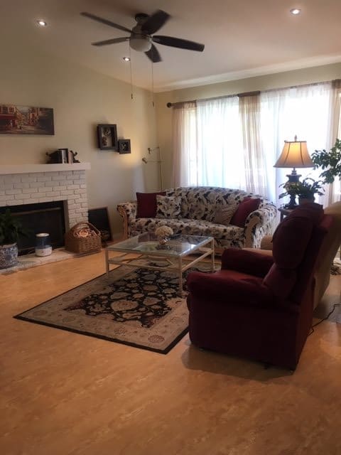 A cozy living room with a sofa and armchair arranged around a fireplace, featuring a decorative rug, a lamp, and large windows with curtains.