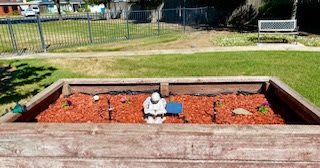 A small garden statue is placed against a wooden border with red mulch, in front of a grassy area and fence.