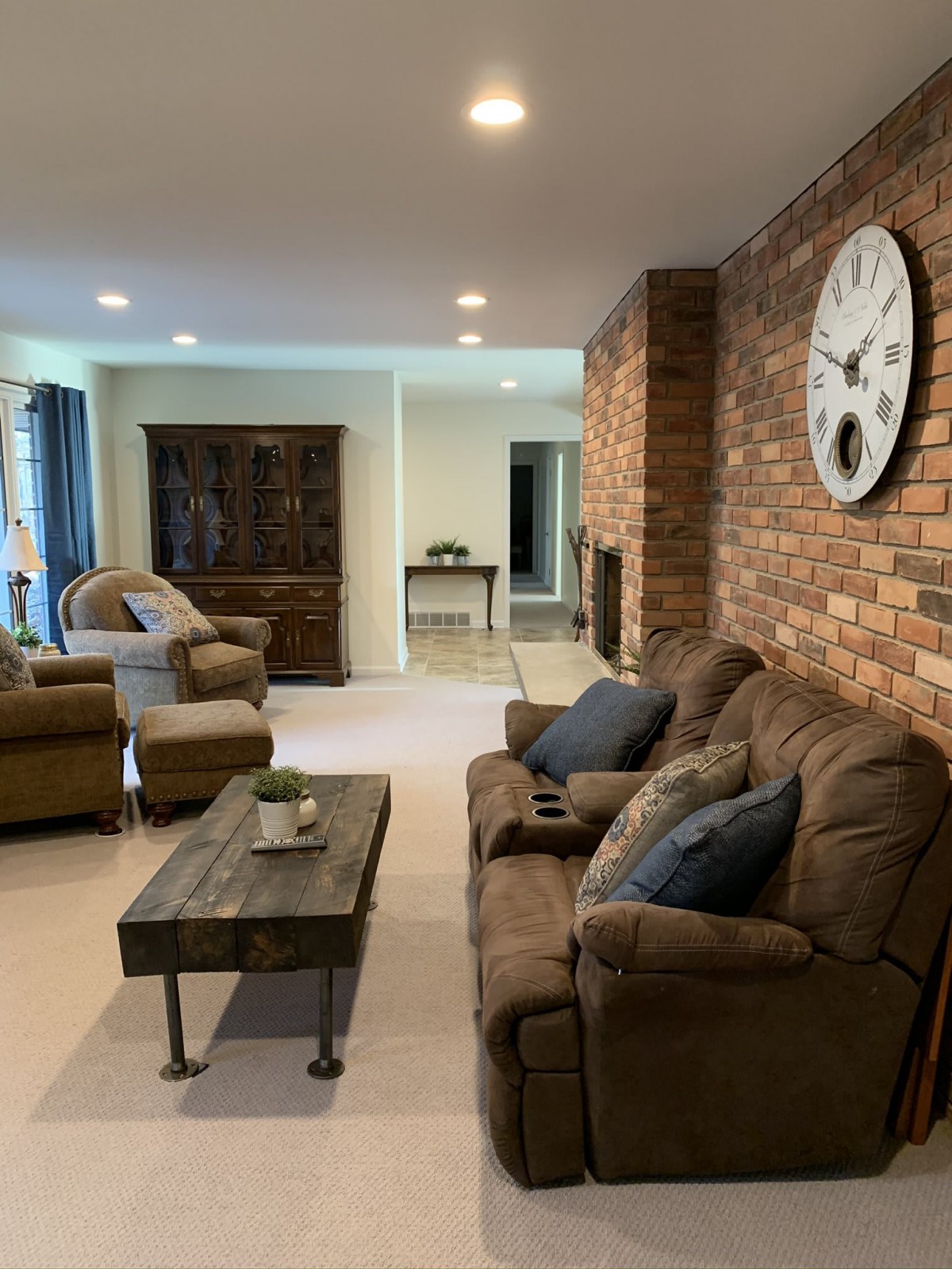 Cozy living room with a brick accent wall, featuring comfortable sofas and armchairs, a rustic coffee table, and a large wall clock.