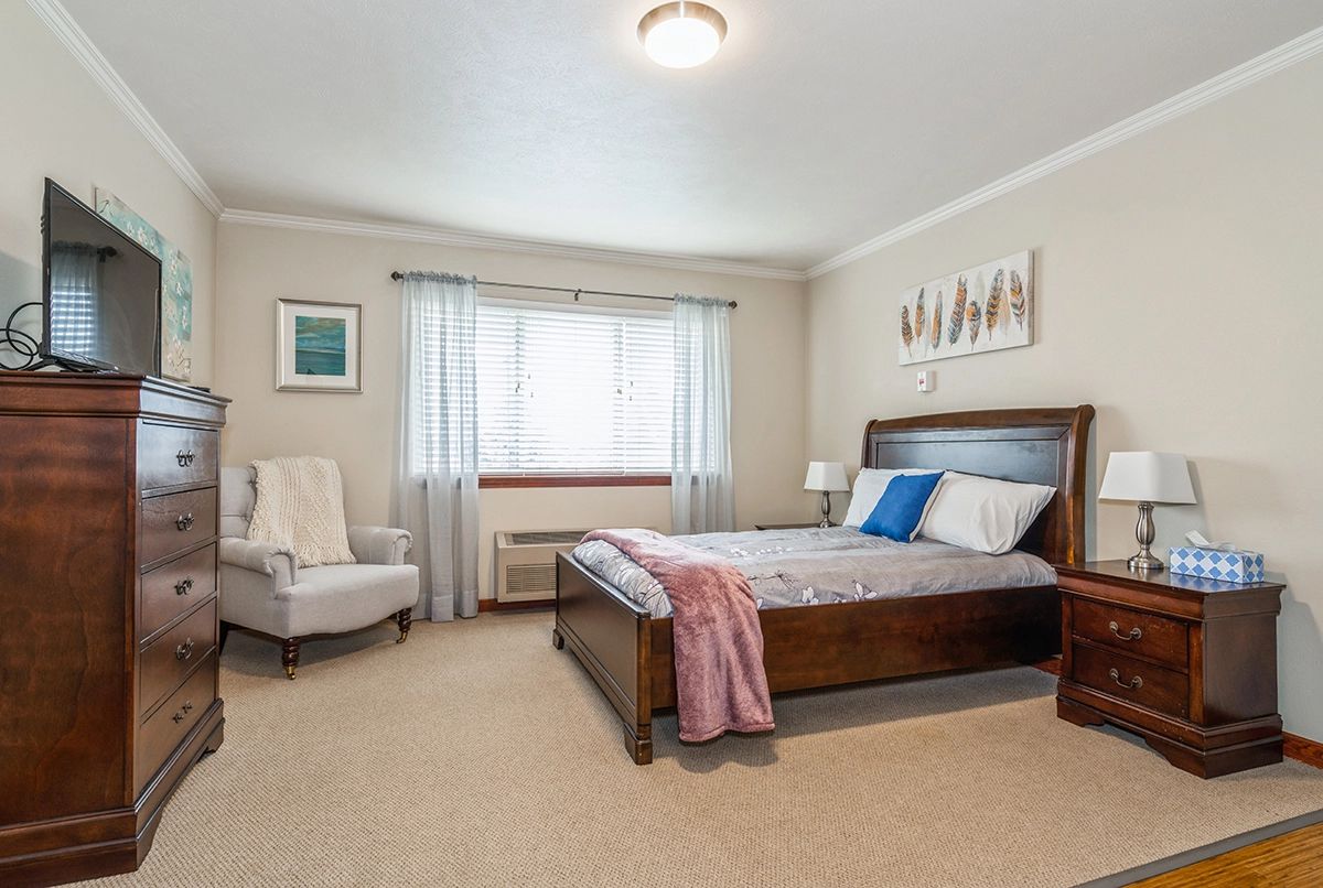 A cozy and well-furnished bedroom with a large bed, an armchair, and bright natural light from the window.