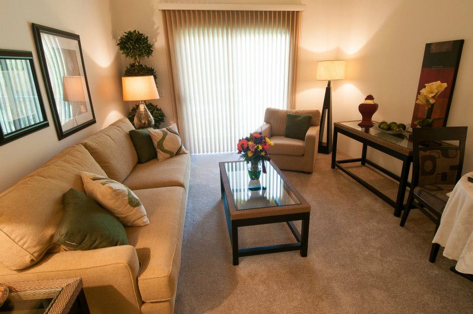 A cozy living room with neutral tones, featuring a sofa, armchair, and a glass-topped coffee table under warm lighting.