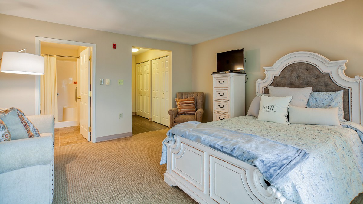 A cozy bedroom featuring a white bed, dresser, television, and armchair.