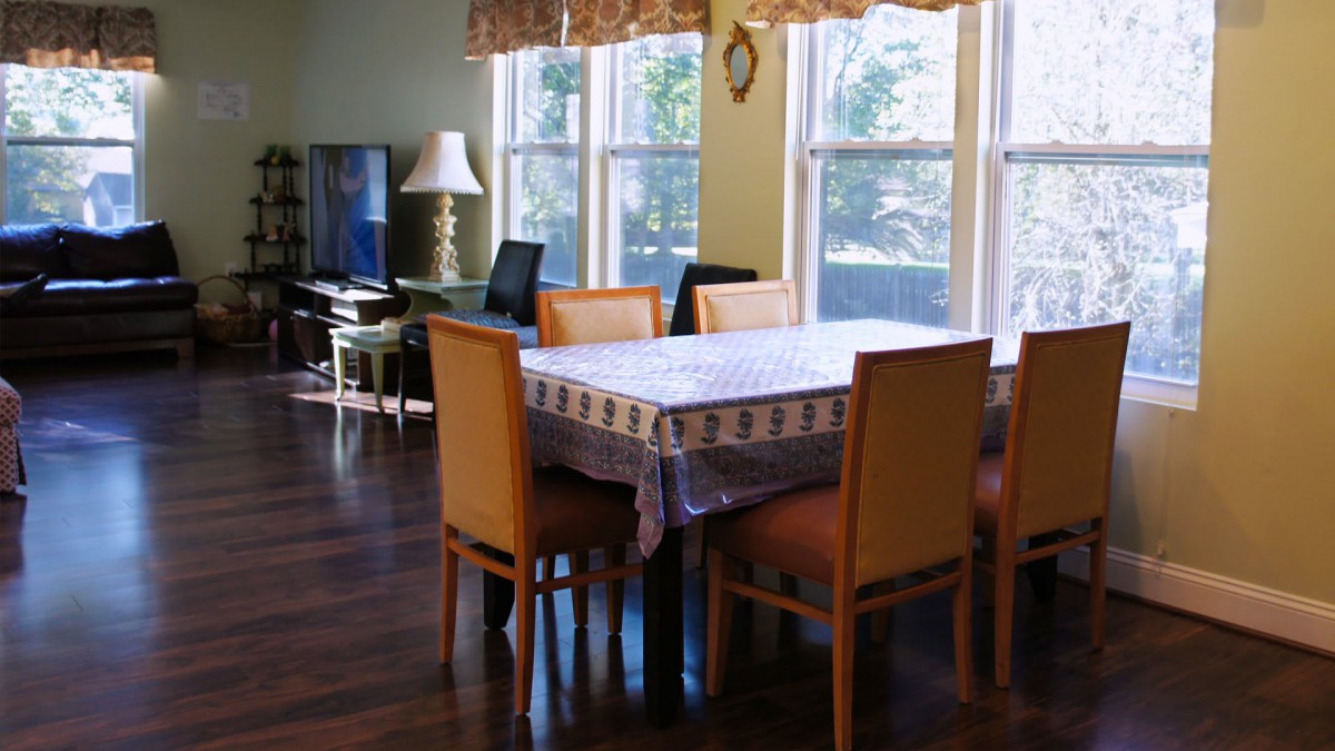 A dining room with a table covered by a cloth, surrounded by chairs and illuminated by natural light through large windows.