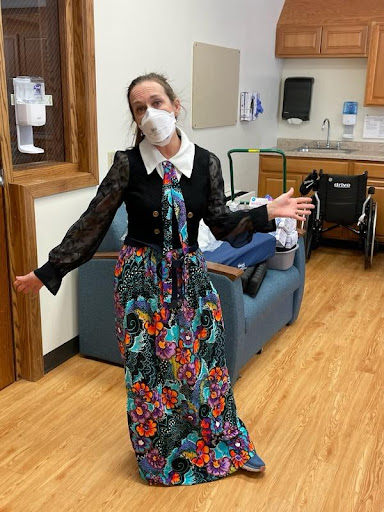 A woman dressed in a colorful floral dress and a black blouse, wearing a mask and standing in a healthcare setting, spreading positivity.