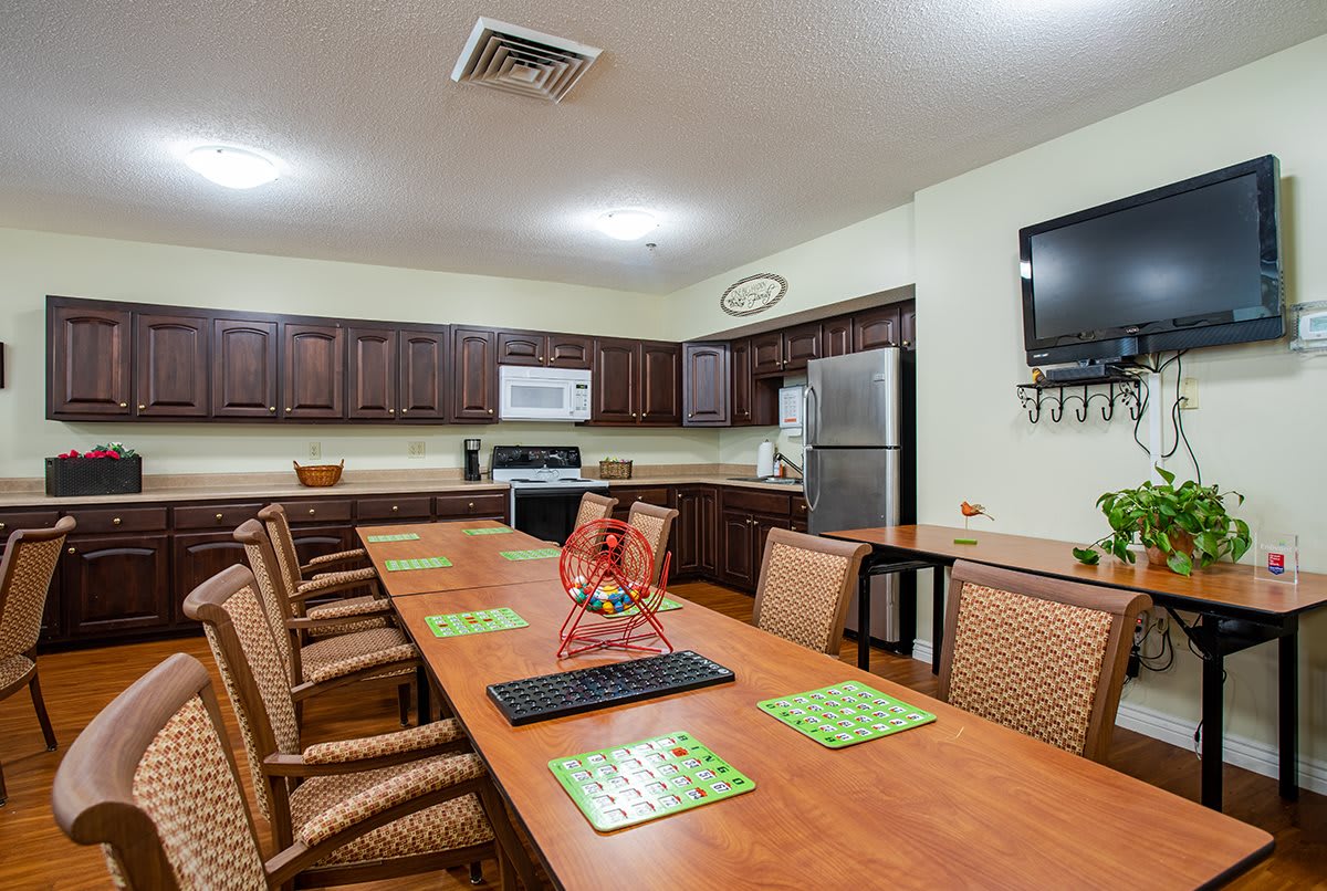 A kitchen area with a long table surrounded by chairs, featuring cabinets, a microwave, refrigerator, and a wall-mounted television.