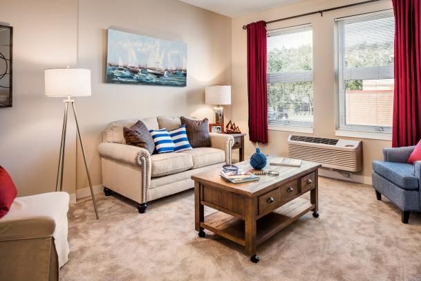 A cozy living room with a beige sofa, decorated with striped and solid pillows, a wooden coffee table, and a painting above the sofa. The room is well-lit by a standing lamp and natural light from the window with red curtains.