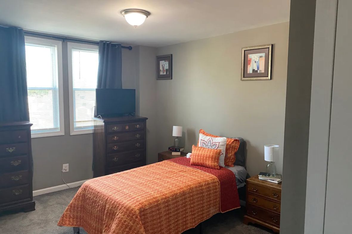 A cozy bedroom with a single bed featuring orange bedding, a dresser with a TV on top, and nightstands with lamps.