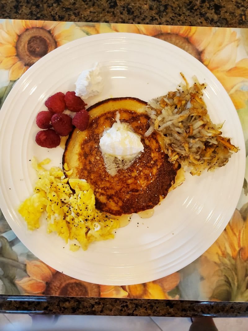 A hearty breakfast plate featuring pancakes with whipped cream, scrambled eggs, hash browns, and fresh raspberries.