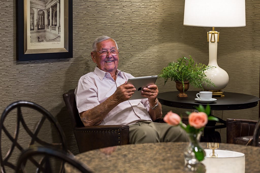 An elderly man sitting in a living room, smiling while using a tablet.
