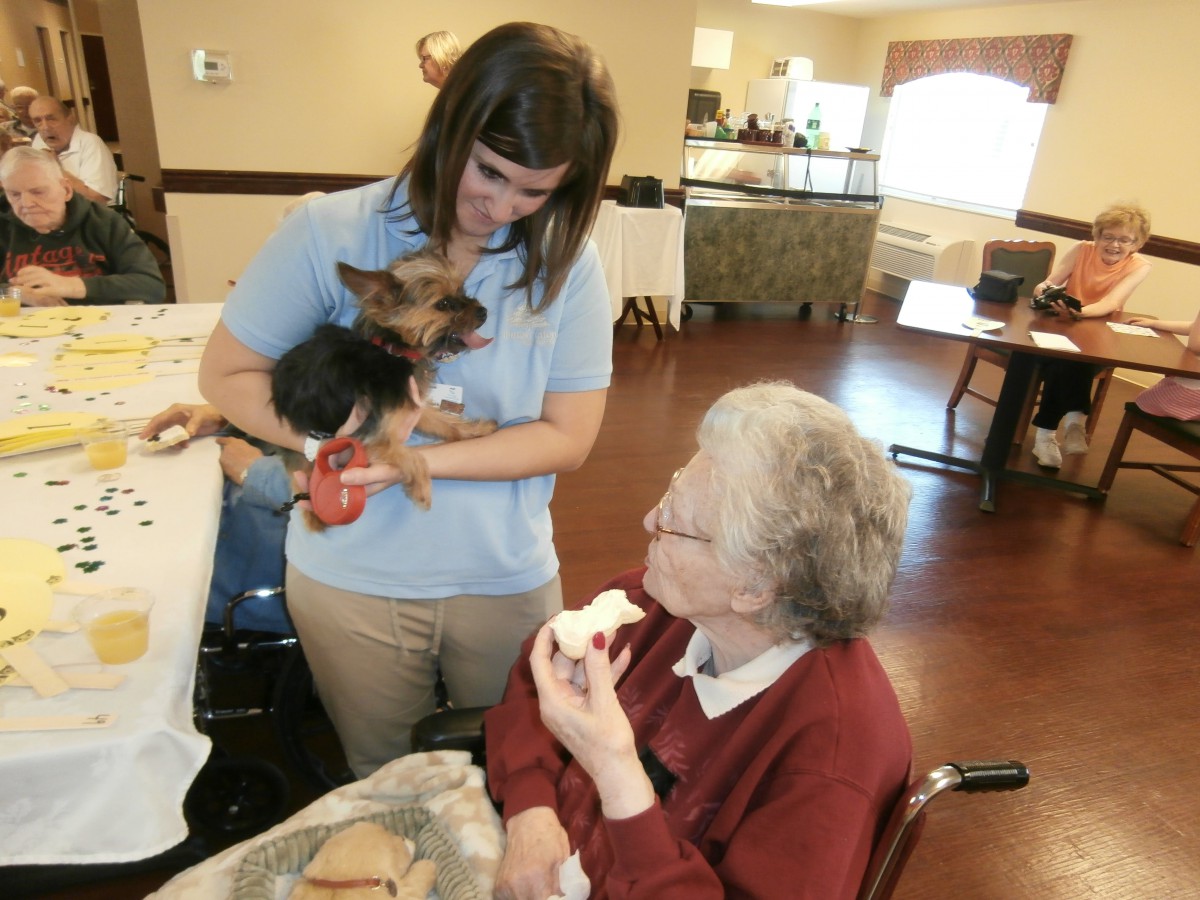 A nurse holds a small dog while interacting with an elderly woman in a care facility.