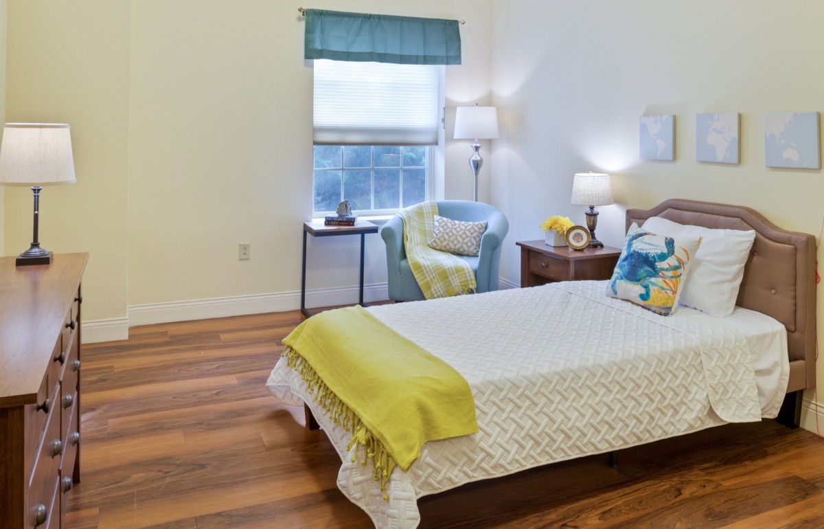 A cozy bedroom with a neatly made bed, armchair, and a window letting in natural light.