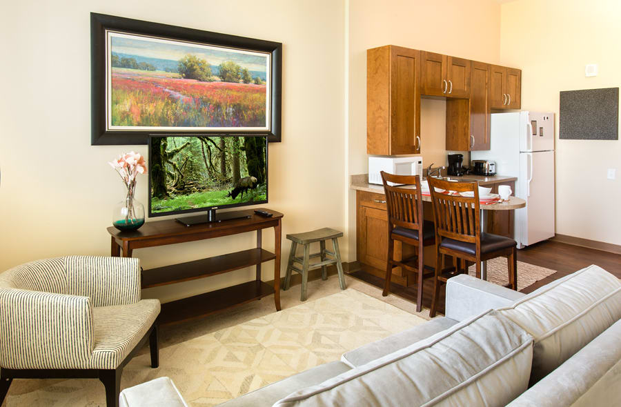 An open layout living room with a television and artwork, adjacent to a kitchen area with wooden cabinets and bar stools.
